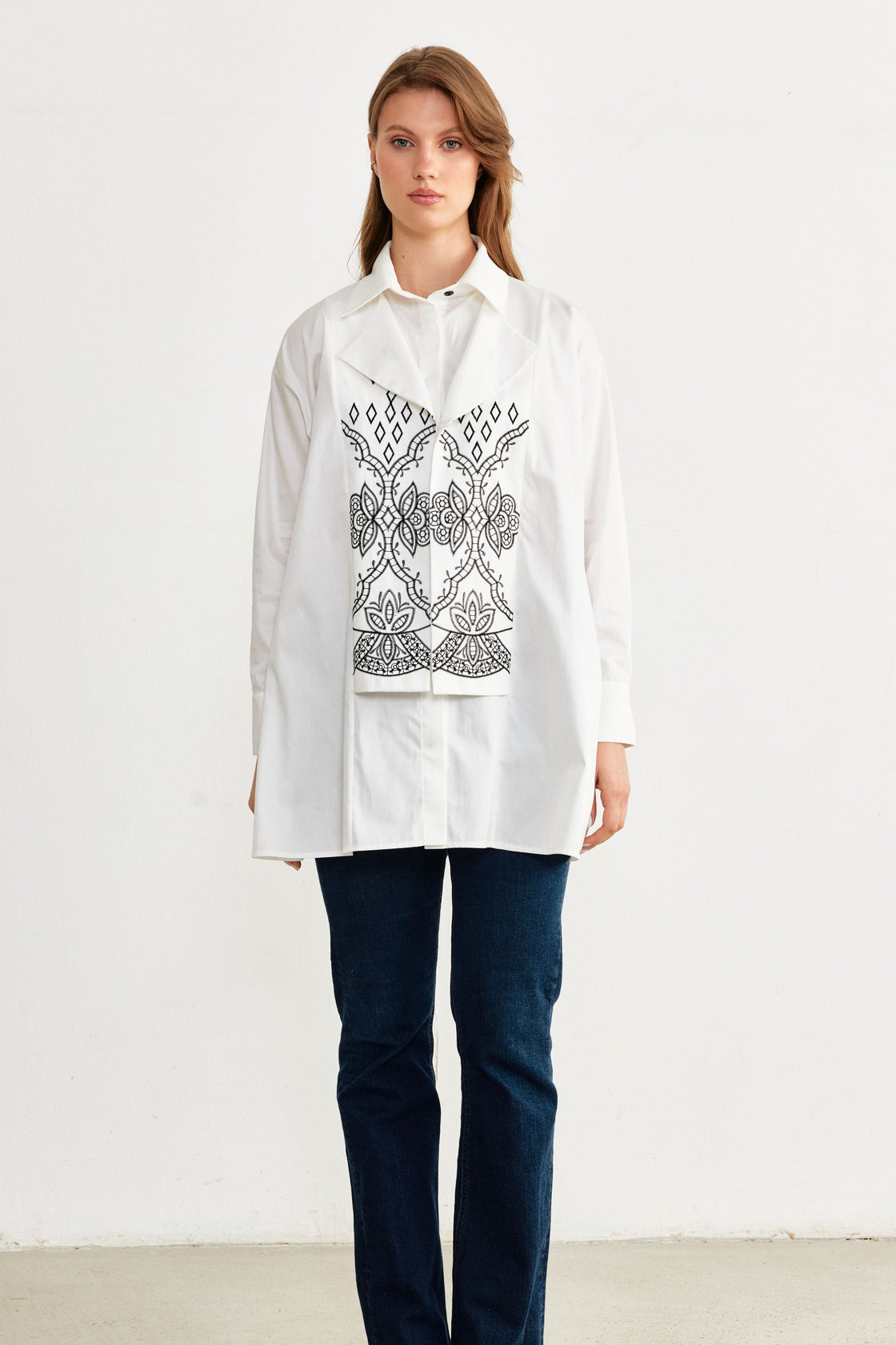 Woman wearing a white shirt with black embroidery and dark jeans on a white background